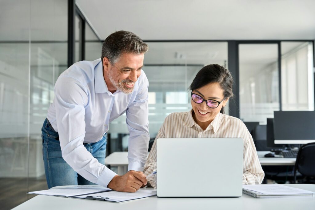 Mid aged manager helping Asian worker working on laptop in office.